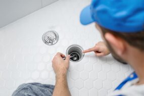 Professional plumber unclogging a shower drain using a specialized tool on a white tiled background. Concept of plumbing maintenance and repair.